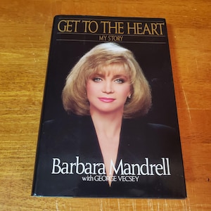 May include: A hardcover book titled "Get to the Heart My Story" by Barbara Mandrell with George Vecsey. The cover features a portrait of Barbara Mandrell with blonde hair and a black background. The book is on a wooden surface.