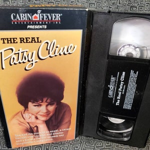 May include: A VHS video cassette tape with a black case and a label that reads "CABIN FEVER ENTERTAINMENT INC PRESENTS THE REAL Patsy Cline 45 Minutes". The front cover of the case features a photo of Patsy Cline.