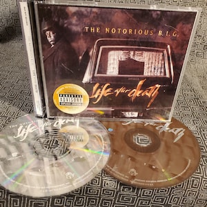 May include: A CD case with a black and white photo of a man in a suit and a hearse. The title of the album is "The Notorious B.I.G. Life After Death". The album cover has a parental advisory sticker.