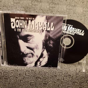 May include: A CD case with a black and white photo of John Mayall on the front. The title of the album is "Silver Tones - The Best of John Mayall & The Bluesbreakers".