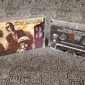 May include: A cassette tape of Traveling Wilburys Vol. 3. The cassette tape is clear plastic with a black label. The label has the band name, album title, and track list. The cassette tape is on a patterned surface.