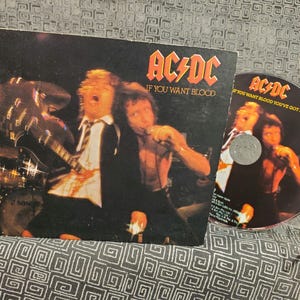 May include: A black and white vinyl record album cover featuring the band AC/DC. The album title is "If You Want Blood". The cover art features a band member playing a guitar and another band member with a microphone.