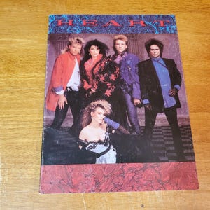 May include: A vintage album cover featuring the band Heart. The cover has a red and blue color scheme with the band members posing in 80s attire. The band's name is displayed in a blue, stylized font at the top of the cover.