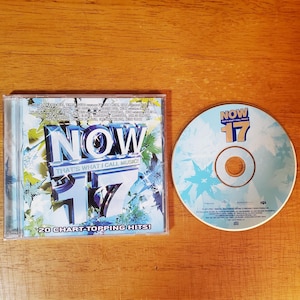 May include: A CD case and disc for "NOW That's What I Call Music! 17". The case has a blue and white design with the text "NOW 17" and "20 Chart-Topping Hits!". The disc is light blue with the text "NOW 17".