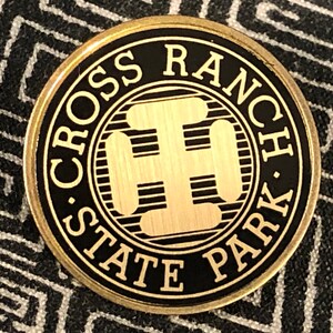 May include: A round, gold-colored metal pin with the words "CROSS RANCH STATE PARK" in white lettering on a black background. The center features a gold-colored design. The pin is set against a patterned background.