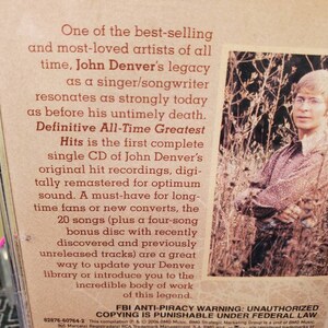 May include: A John Denver CD case back cover featuring a sepia-toned photograph of the artist standing in tall grass. The text describes the Definitive All-Time Greatest Hits collection, including 20 songs and a four-song bonus disc.
