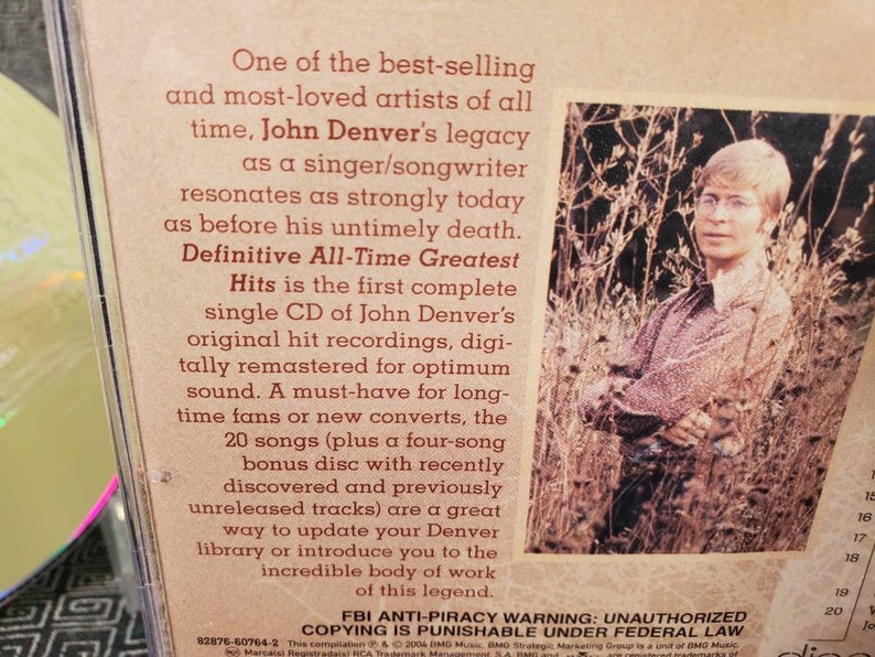 May include: A John Denver CD case back cover featuring a sepia-toned photograph of the artist standing in tall grass. The text describes the Definitive All-Time Greatest Hits collection, including 20 songs and a four-song bonus disc.