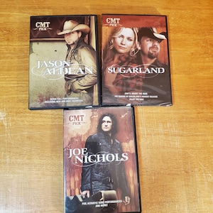 May include: Three CMT Pick DVDs featuring country music artists. The DVDs include Jason Aldean, Sugarland, and Joe Nichols. Each DVD case has a photo of the artist and the name of the artist. The DVDs are on a wooden surface.