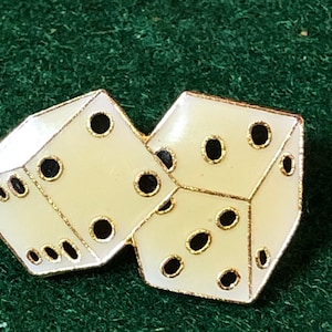 May include: A pair of dice-shaped pins. Each die is off-white with black dots and gold trim. The dice are interlocked, suggesting a game or gambling theme. The background is a dark green.