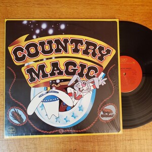 May include: A vinyl record album titled Country Magic featuring a cartoon rabbit emerging from a magician's hat on the cover. The artwork includes a yellow border, a black background with white stars, and small illustrations of corn and a pig.