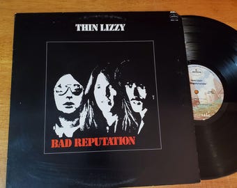 May include: A black vinyl record album by Thin Lizzy, titled "Bad Reputation." The album cover features a black and white image of the band members. The album is partially visible, with the record sleeve and the vinyl record itself.