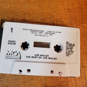 K&ouml;nnte beinhalten: Eine wei&szlig;e Kassette mit dem Album "The Best of Joe Walsh". Das Band hat schwarze kreisf&ouml;rmige Naben und der Text "ROCKY MOUNTAIN WAY" und "JOE WALSH" sind sichtbar. Die Kassette liegt auf einer Holzoberfl&auml;che.