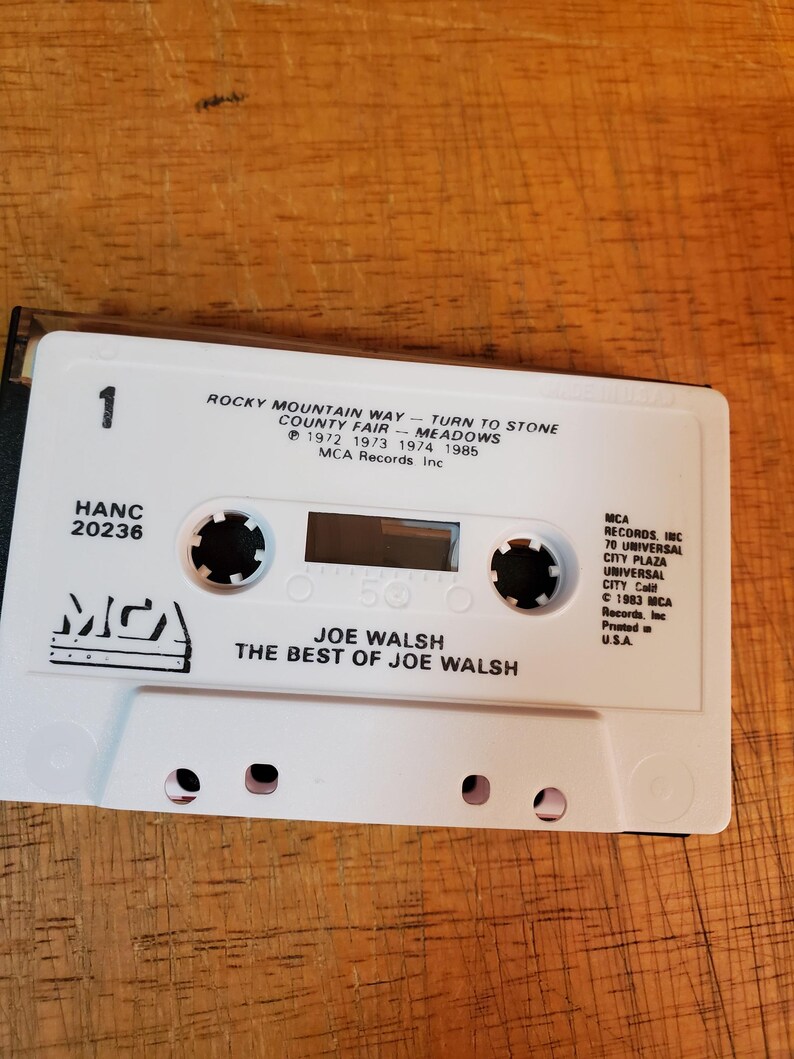 K&ouml;nnte beinhalten: Eine wei&szlig;e Kassette mit dem Album "The Best of Joe Walsh". Das Band hat schwarze kreisf&ouml;rmige Naben und der Text "ROCKY MOUNTAIN WAY" und "JOE WALSH" sind sichtbar. Die Kassette liegt auf einer Holzoberfl&auml;che.