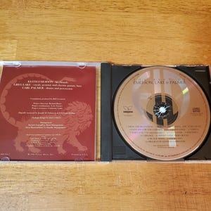 May include: Open CD case with a disc featuring the text "The Best of Emerson, Lake & Palmer." The left side of the case displays text including band member names and a lion graphic. The disc has a silver surface with black text.
