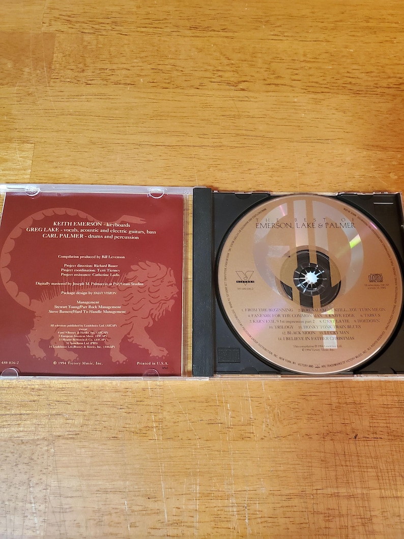 May include: Open CD case with a disc featuring the text "The Best of Emerson, Lake & Palmer." The left side of the case displays text including band member names and a lion graphic. The disc has a silver surface with black text.
