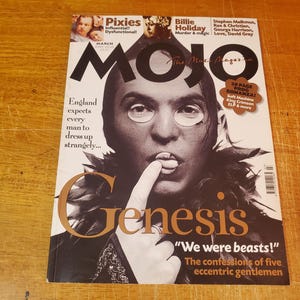 May include: A magazine cover featuring a black and white photo of a man with a feathered collar. The magazine is titled "MOJO The Music Magazine" and the cover story is "Genesis: "We were beasts!" The confessions of five eccentric gentlemen."