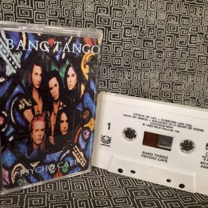 May include: A white cassette tape with a black and red cassette tape inside. The cassette tape is labeled "Bang Tango Psycho Cafe" and features the song titles "Attack of Life", "Someone Like You", "Wrap My Wings", "Breaking Up a Heart of Stone", and "Shotgun Man".