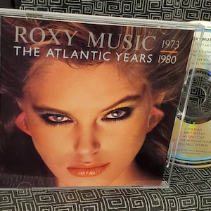 May include: A CD case with a close-up image of a woman with heavy eye makeup. The case is for a compilation album of Roxy Music's music from 1973 to 1980, titled "The Atlantic Years 1980".