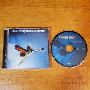 May include: A music CD titled Uriah Heep High and Mighty in a clear plastic jewel case alongside the matching disc. The cover art features a stylized airplane against a blue sky with white clouds. The disc displays the same airplane graphic.
