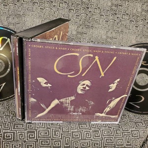May include: A CD set featuring Crosby, Stills & Nash. The album cover is a deep purple with gold lettering and images of the band members. Two CDs are visible, one labeled "Disc 1" and the other "Disc 2". The album title is "Carry On".