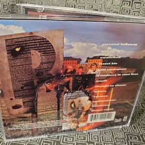 以下が含まれることがあります： 画像とテキストのコラージュが施された音楽CDの背面。テキストには、「personal holloway」、「greedy ay」、「swallowed」、「insect kin」、「cold contagious」、「tendency to start fires」、「mouth」、「straight no chaser」、「history」、「synapse」、「communicator」、「home driver」、「distant voices」などの言葉が含まれています。