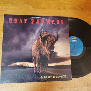 May include: A vinyl record album by Beat Farmers. The album cover features a brown bison standing on a road with a cloudy sky background. The album title is "The Pursuit of Happiness."