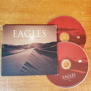 May include: The album "Long Road Out of Eden" by the Eagles. The album cover features a desert landscape in sepia tones. Two red CDs are visible. The text "EAGLES" is displayed on the album cover and one of the CDs.