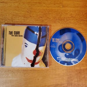 May include: A CD case and disc for The Cure's album "Wild Mood Swings." The case features a close-up of a person's face with a clown-like design. The CD is blue with white text.