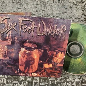 May include: A CD and its case for the album "The  Haunted  Life" by the death metal band Six Feet Under. The album cover features a dark, grungy aesthetic with jars filled with various objects.