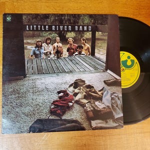 May include: An album cover for the Little River Band. The cover features a group of men standing on a wooden structure. The album is next to a black vinyl record with a yellow label. Clothing and a hat are on the ground.