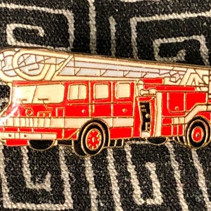 May include: A red and white enamel pin of a fire truck with a ladder. The truck has a white cab and a red body with black details. The ladder is white and has black rungs.