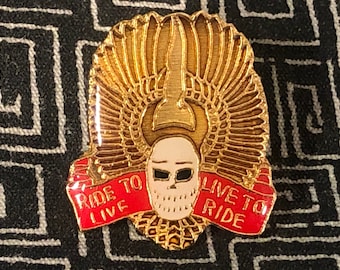 May include: Gold-coloured pin featuring a skull wearing a winged helmet and a red ribbon with the text "Ride to Live Live to Ride".