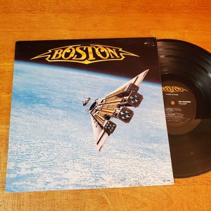 May include: Album cover for Boston's "Third Stage" vinyl record. The cover features a spaceship design against a blue and white background. The band's name, "BOSTON", is in gold lettering. A black vinyl record is visible.