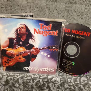 May include: A CD and a CD case featuring Ted Nugent. The CD case is black with a red and white title, "Ted Nugent: Motor City Madness". The CD is black with a red and white title, "Ted Nugent: Motor City Madness".