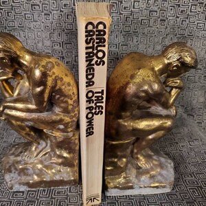 May include: A pair of gold-toned bookends shaped like the "Thinker" statue, holding up a book titled "Tales of Power" by Carlos Castaneda.
