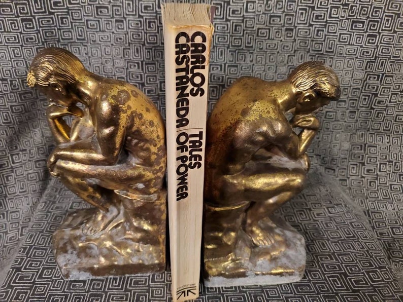 May include: A pair of gold-toned bookends shaped like the "Thinker" statue, holding up a book titled "Tales of Power" by Carlos Castaneda.