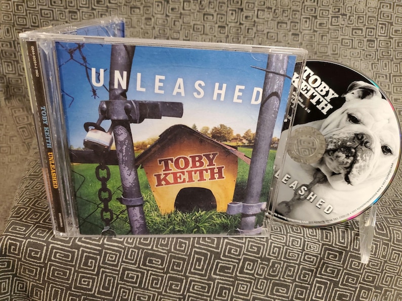 May include: A Toby Keith Unleashed music CD in a clear plastic jewel case. The cover art features a yellow doghouse behind a chain-link fence with a padlock. The disc itself is printed with a black and white image of a bulldog and the text Toby Keith.