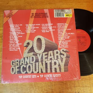 May include: A 20 Grand Years of Country vinyl record album cover in red with white text and a wooden block graphic. The cover lists 20 songs and artists, including Willie Nelson and Johnny Cash, with a barcode and a black vinyl record visible.