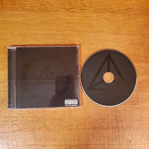 May include: A black CD case and a matching black compact disc. The case cover features a faint triangular symbol and a Parental Advisory Explicit Content label. The disc also displays a dark triangular graphic centered on its surface.