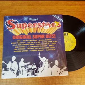 May include: A Ronco Presents Superstars Original Super Hits vinyl record album cover and a black vinyl record. The cover is dark blue with yellow text, featuring a list of artists including Kiss, Donna Summer, and Abba, and a white band silhouette.