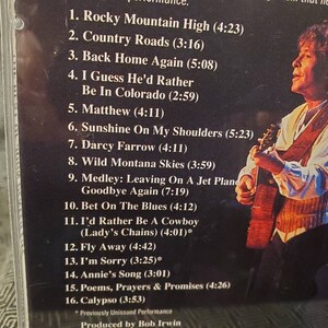 May include: A black and white CD case with a photo of a man playing a guitar. The case lists the songs on the CD, including "Rocky Mountain High", "Country Roads", and "Back Home Again".