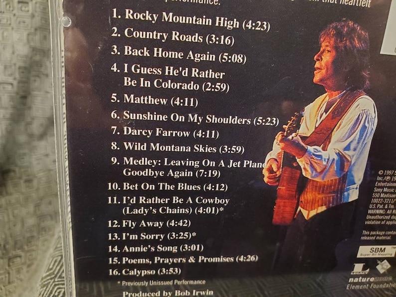 May include: A black and white CD case with a photo of a man playing a guitar. The case lists the songs on the CD, including "Rocky Mountain High", "Country Roads", and "Back Home Again".