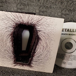 May include: A white CD case with a black and white image of a coffin inside a magnetic field. The CD case is for the Metallica album "Death Magnetic".