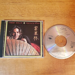 May include: A Tommy Bolin Private Eyes music CD in a clear plastic case with a cover featuring a person holding a large fan and Japanese text. The silver disc displays the album title, track list, Columbia logo, and compact disc digital audio symbol.