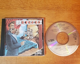 May include: A CD of "The Best of Kansas" album. The album cover features a man in a suit, a ship, and the band's name. The CD is next to the case, with the tracklist visible. The CD is on a wooden surface.