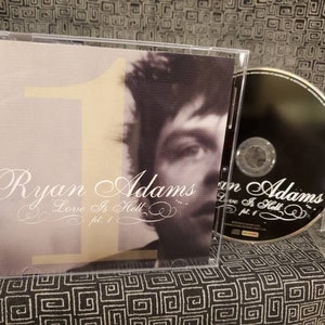 May include: A CD case with a black CD inside. The front cover features a black and white photo of Ryan Adams with the text "Ryan Adams Love Is Hell pt. 1" in white.