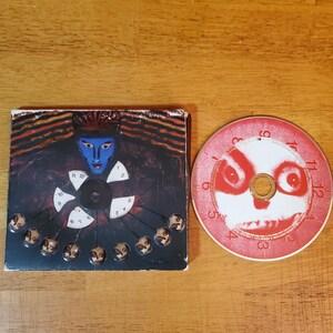 May include: A CD case and a CD are displayed on a wooden surface. The CD case features a blue-faced figure with a clock design. The CD has a red and white design with a face-like pattern and numbers.