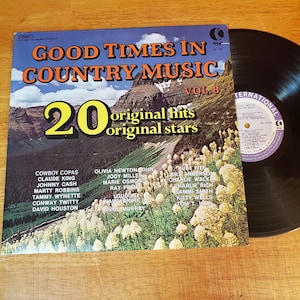 May include: A vinyl record album titled Good Times In Country Music Vol. 8, featuring a mountain landscape with white wildflowers. The cover lists 20 original hits and stars including Johnny Cash and Tammy Wynette. A black vinyl disc is partially visible.