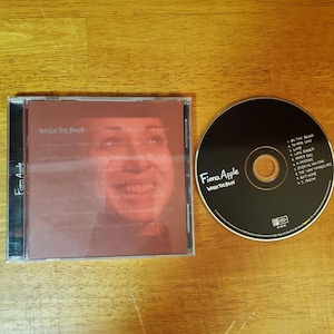 May include: A CD case and disc featuring Fiona Apple's album "When the Pawn...". The CD case has a reddish-brown cover with a blurred image of a person's face. The disc is black with white text, including the artist's name and album title.
