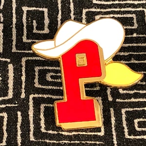 May include: A red letter "P" with a gold outline and a white cowboy hat with a yellow brim. The pin is on a black and white patterned fabric.